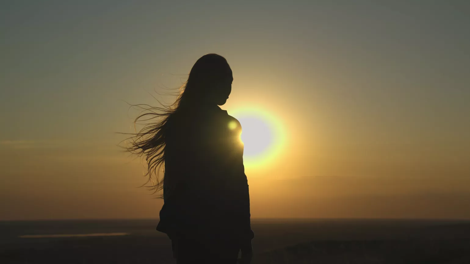 Young girl looks at sunset silhouette, hair blowing in the wind