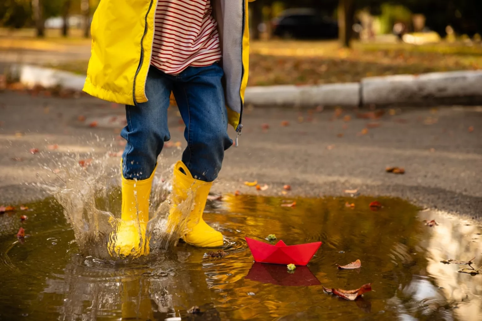 Kid with yellow raincoat and boots, splashing in a puddle with a red paper boat