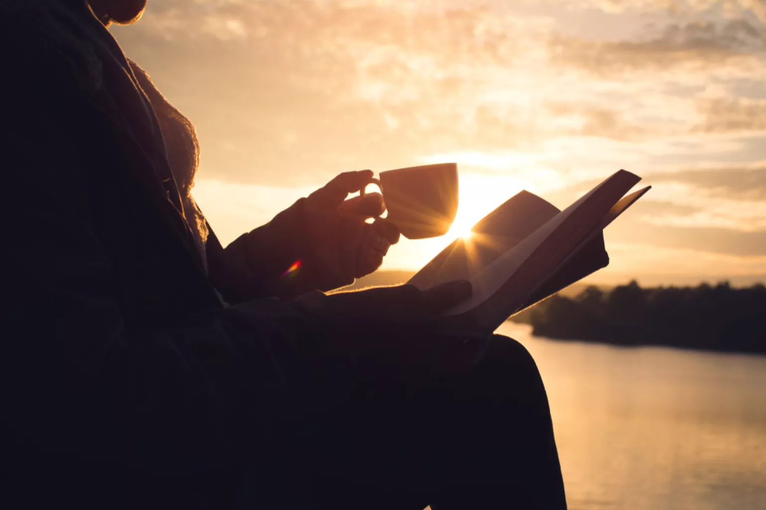 Silhouette of a woman reading a book sitting on the rock near lake waiting for sunset 