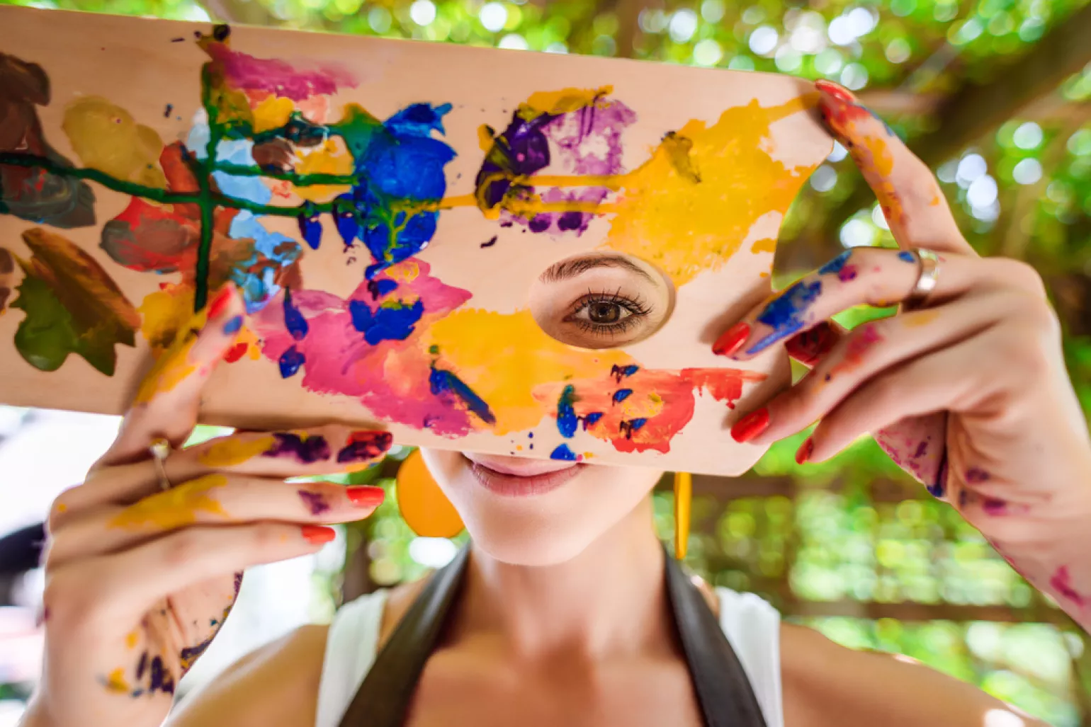 Playful portrait of a young female artist painter covered in paint, looking and smiling at camera through her painter's palette.