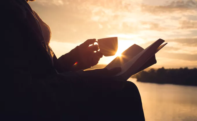 Silhouette of a woman reading a book sitting on the rock near lake waiting for sunset 